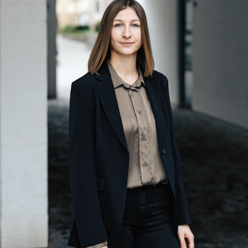 Junge Frau mit Bluse und Jacket, die draußen in einem Hausflur steht.