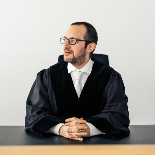 Richter in Robe, der mit verschränkten Händen am Tisch sitzt und zur Seite blickt