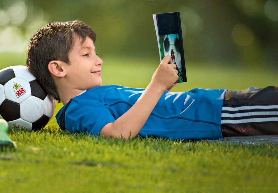Das Foto eines Jungen, der i einem Buch liest. Dabei liegt er auf dem Rücken auf einem Fußballfeld, den Kopf auf einen Fußball aufgestützt.