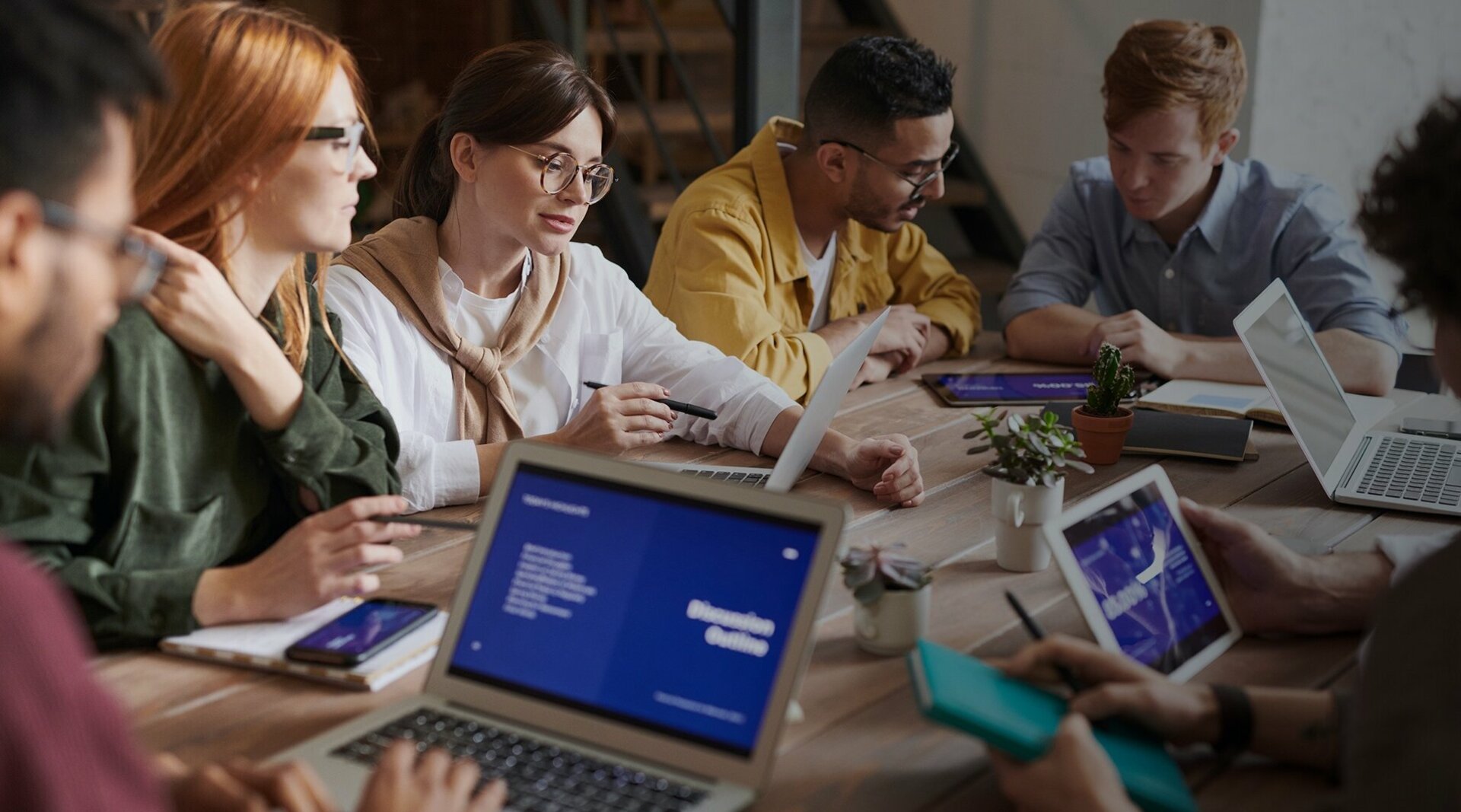 Sieben junge Erwachsene sitzen um einen Tisch mit ihren Laptops und haben ein Meeting.