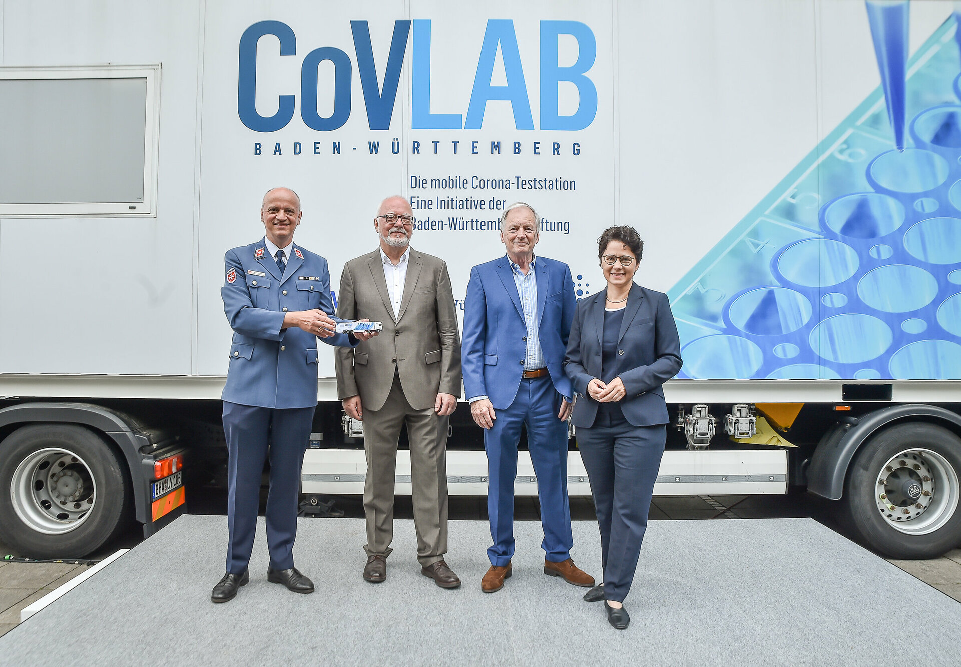 Klaus Weber, Prof. Dr. med. Michael Neumaier, Christoph Dahl und Marion Gentges stehen vor dem CovLAB. Klaus Weber hält ein Modell des CovLAB-Trucks in der Hand.