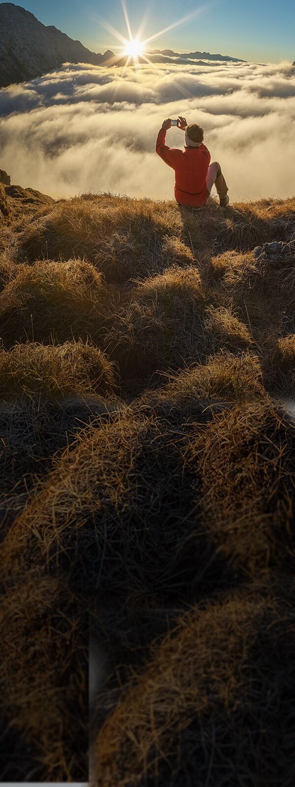 Man sieht die Rückansicht eines Mannes. Er sitzt auf einem Berg und fotografiert den Sonnenaufgang. Vor ihm ziehen die Wolken vorbei.