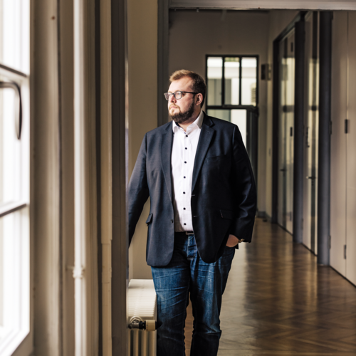 Mann mit Jeans, Jacket, Bart und Brille, der aus dem Fenster eines Verwaltungsgebäudes schaut