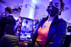 Eine Frau mit Mund-Nasen-Schutz hält ihr Smartphone in die Kamera. Darauf sieht man ein Foto der Veranstaltung Globa Afterwork als Instagram-Post