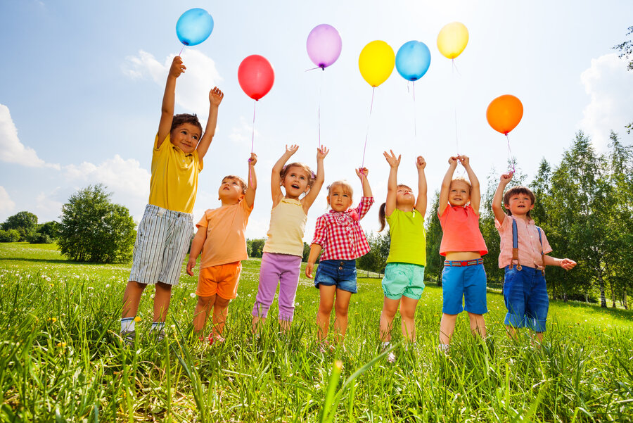 Sieben Kinder stehen auf einer grünen Wiese und spielen mit Luftballons. 
